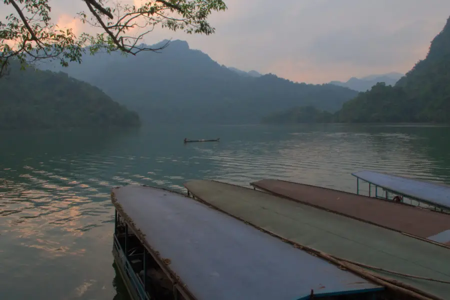 Tour boats floating at the edge of a pristine lake in Vietnam at sundown. A man is fishing in the background.