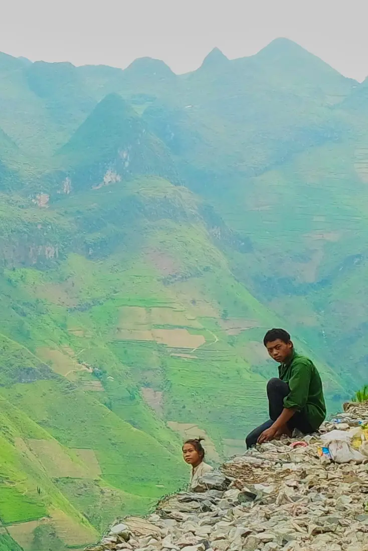 A man sitting on the edge of a cliff road with mountains behind him at Ma Pi Leng pass, Ha Giang